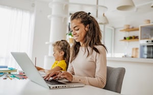 A woman sits at a table working on a laptop, completing online coursework at home, while a young child beside her colors with pencils in a bright, modern kitchen.