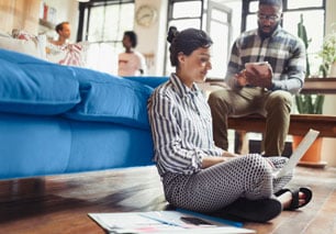 Female sitting on the floor with her back against a couch while browsing degree programs on her laptop
