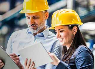 Two engineering technologists wearing yellow hard hats review a control system on their tablets.
