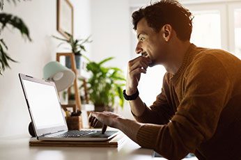 Young man thoughtfully using laptop at bright home office desk
