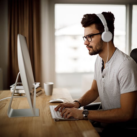 Young man focused typing at desktop computer wearing headphones in bright home office