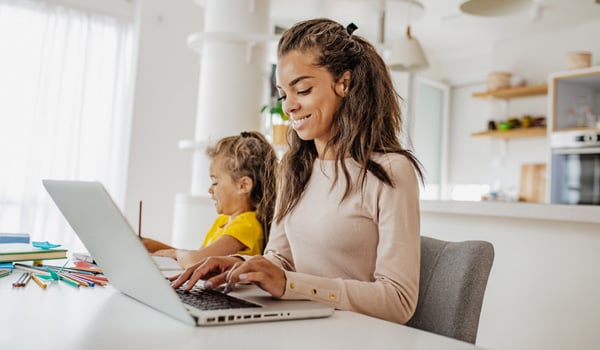 A woman sits at a table working on a laptop, completing online coursework at home, while a young child beside her colors with pencils in a bright, modern kitchen.