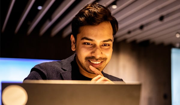 A smiling man sits indoors working on a laptop, resting his hand on his chin in a modern office setting.