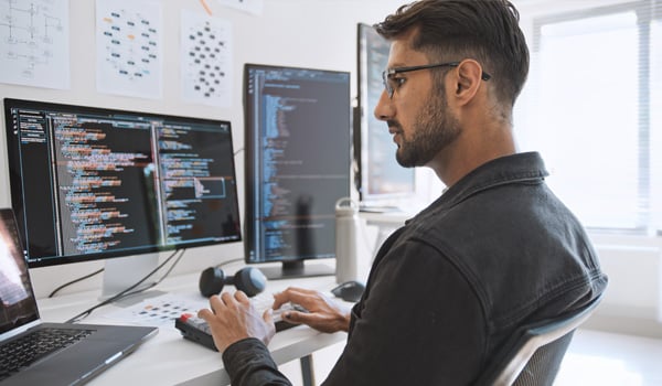 A man wearing glasses works at a desk with dual monitors displaying lines of computer code, typing on a keyboard in a modern office while a woman stands in the background.