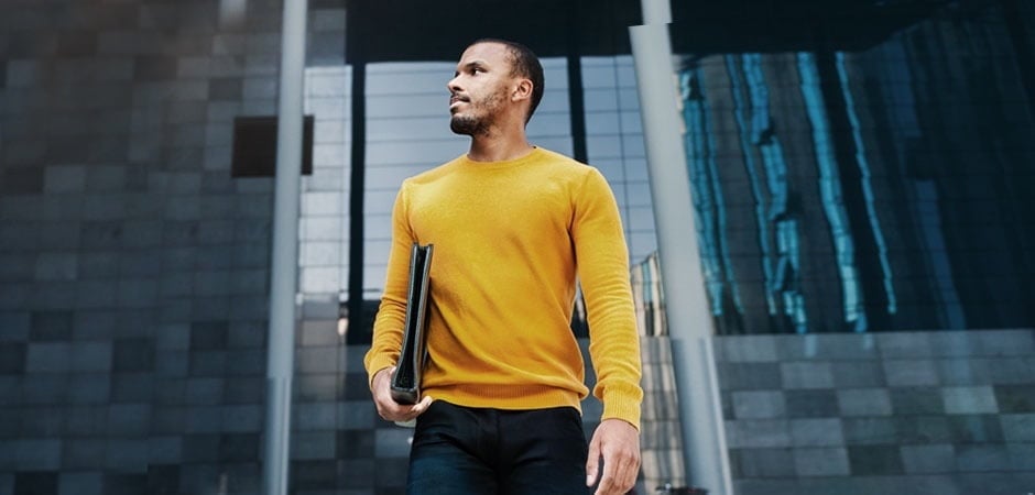 Black man in a yellow sweater holding a notebook under his arm and standing outside of an urban office building after earning his bachelor’s degree.